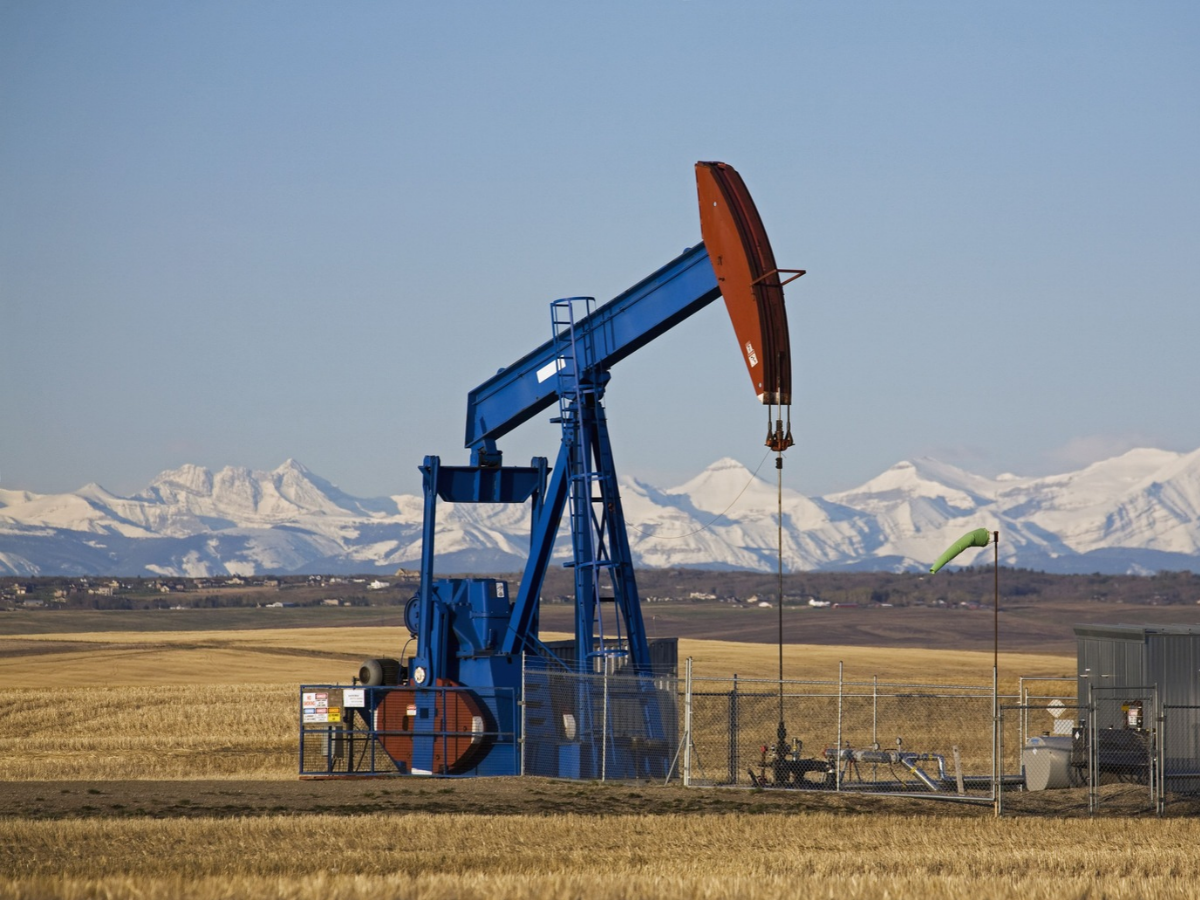 An oil pumpjack stands in a field in Alberta, as snowcapped mountains stand in the background.