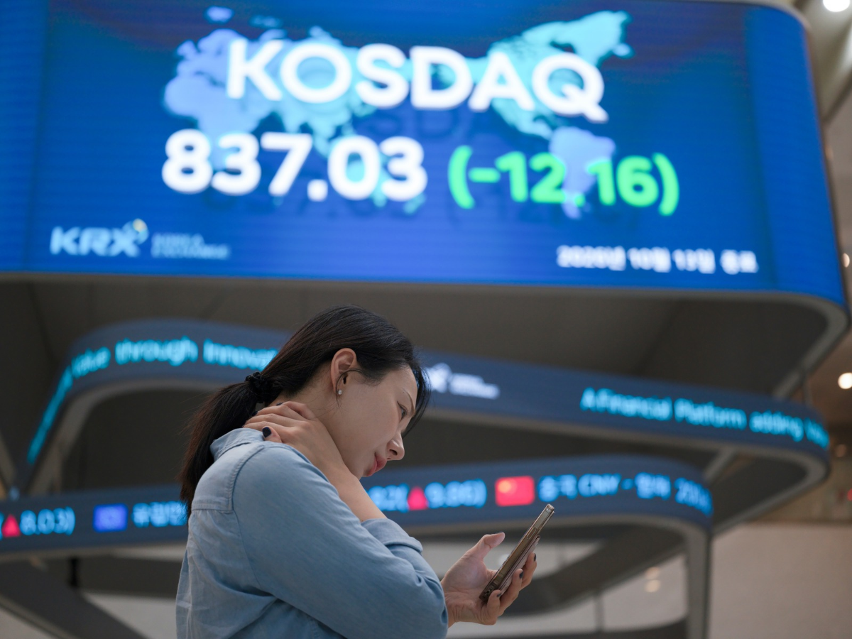 A Korea Exchange (KRX) employee looks at her smartphone in front of a board displaying the closing KOSDAQ index at 837.03 (-12.16) in Yeouido, Seoul, South Korea, on October 13, 2026.