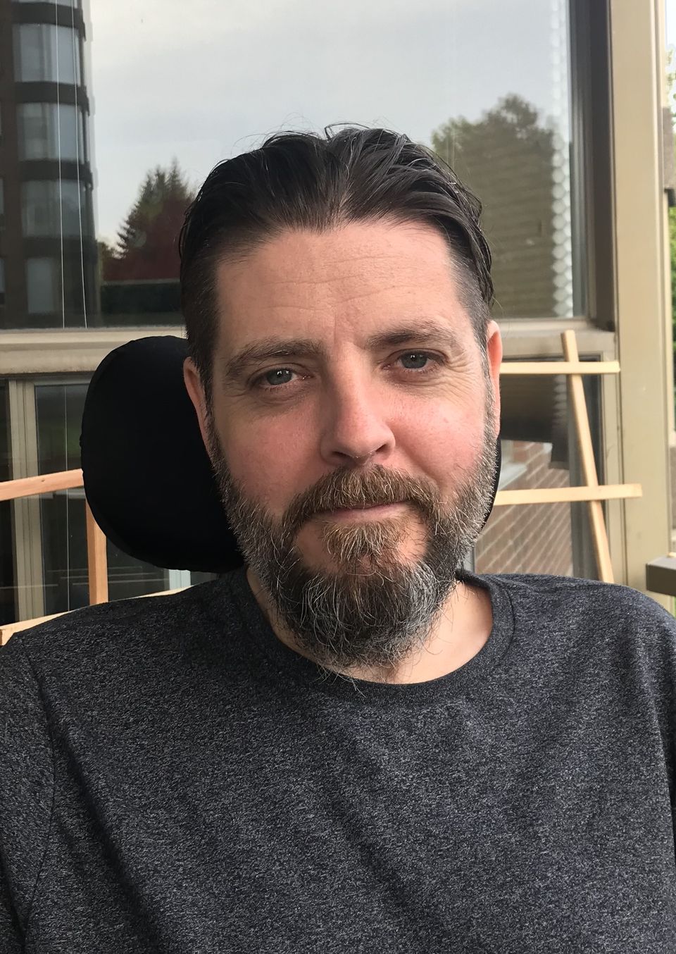 Adam Welburn-Ross, a patient with ALS, wears a grey shirt and looks into the camera. He has a salt-and-pepper beard with brown hair combed back.