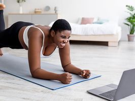 Plus size Afro woman working out at home with online personal trainer, using laptop, standing in elbow plank pose