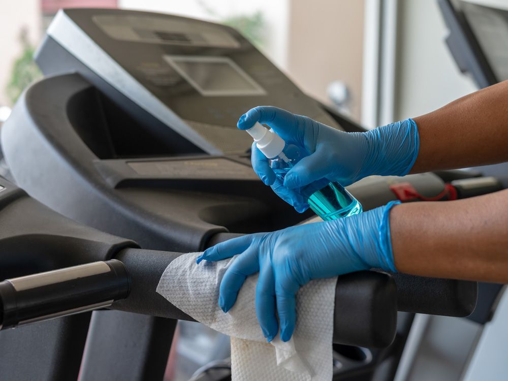 staff using wet wipes and a sanitizer from the bottle to cleana treadmill at a gym.