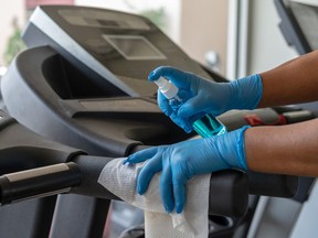 Staff using wet wipes and a sanitizer from the bottle to cleana treadmill at a gym.