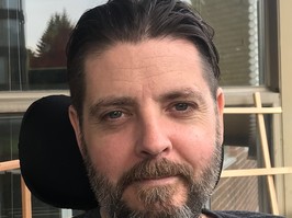 Adam Welburn-Ross, a patient with ALS, wears a grey shirt and looks into the camera. He has a salt-and-pepper beard with brown hair combed back.