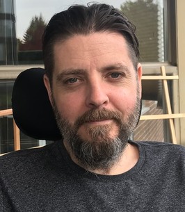 adam welburn-ross, a patient with als, wears a grey shirt and looks into the camera. he has a salt-and-pepper beard with brown hair combed back.