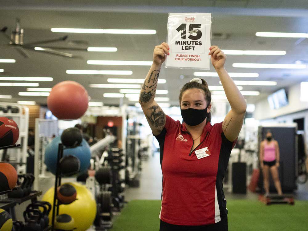 A staff member wearing a protective mask holds up a sign indicating the remaining time for members to complete their workouts before they must leave to allow staff to complete a 30- minute cleaning regimen at a GoodLife Fitness Centers Inc. gym in Ottawa in summer 2020.