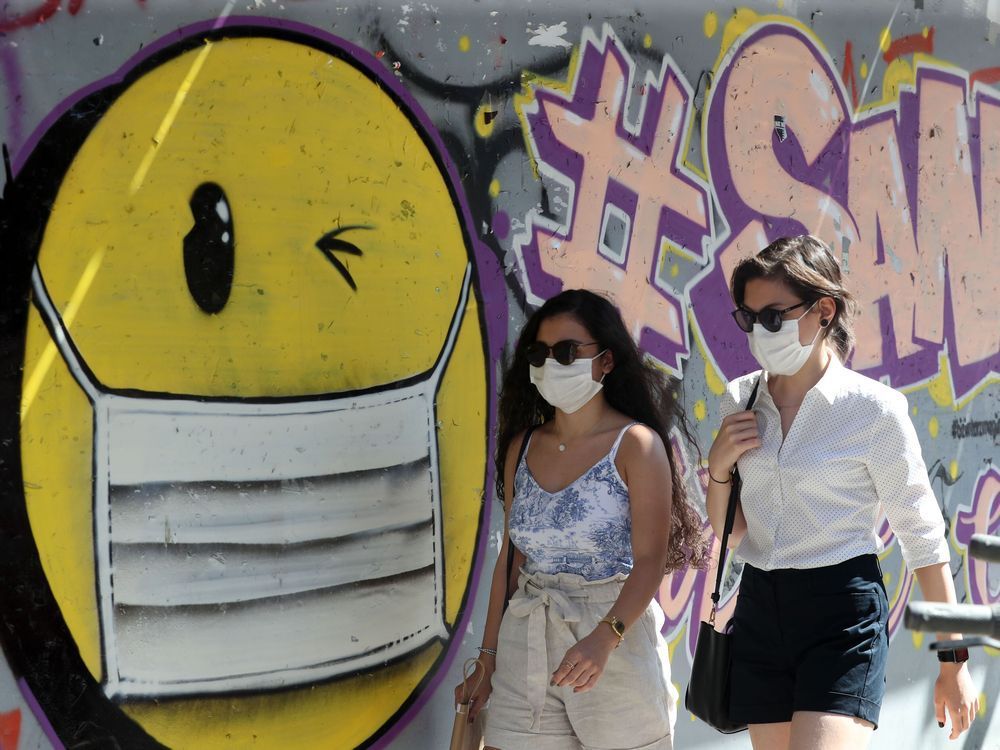 file: women wearing face masks walks past graffiti depicting a smiley face with a face mask.