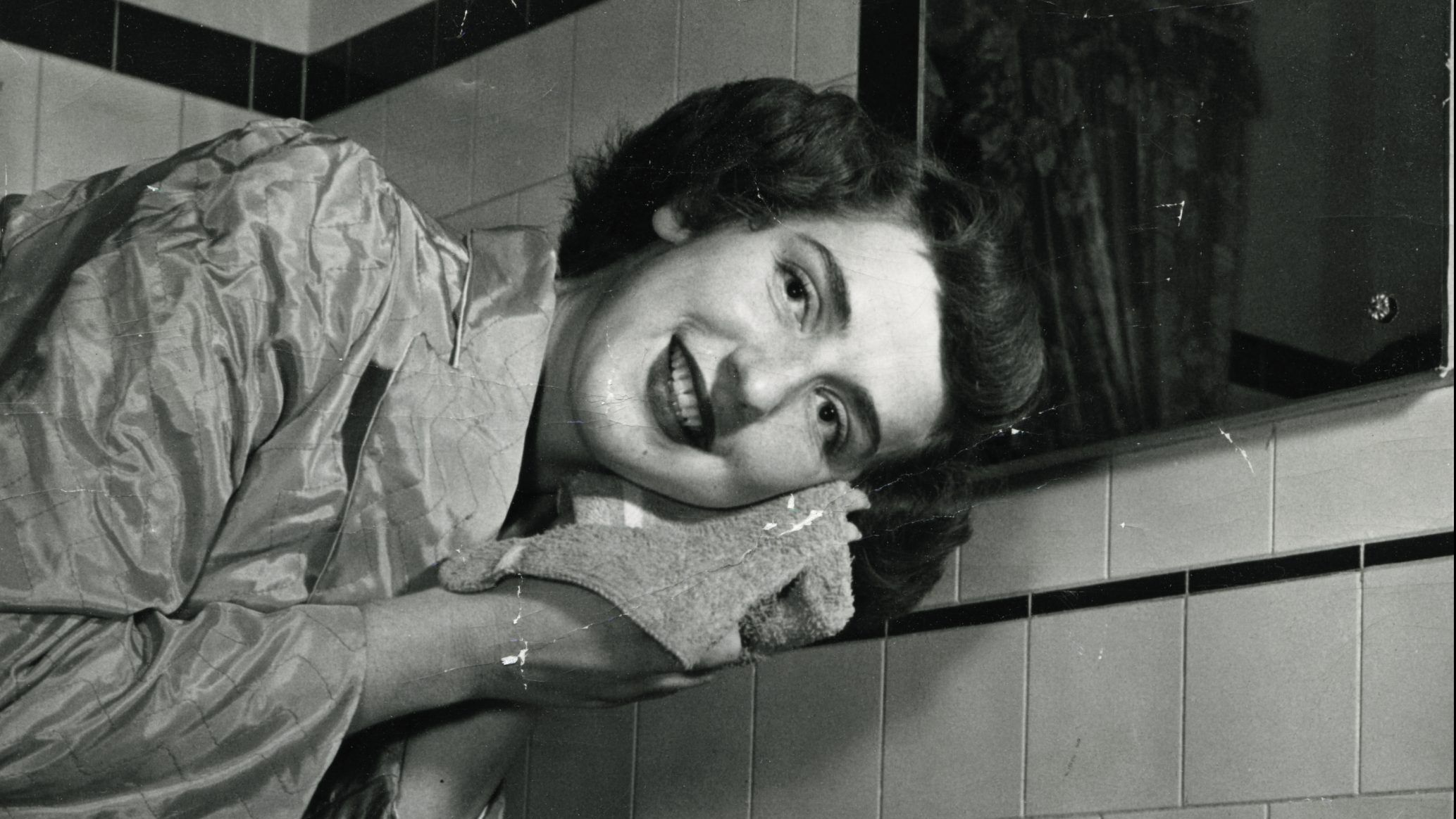 This is a photo taken in the 1940's. A young woman is washing her face in her satin robe. Her face is turned towards the camera, and she is smiling.