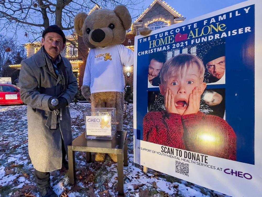 shawn turcotte poses with the cheo bear in front of the family's house, which has been decorated with a home alone theme with a goal of raising $100k for cheo mental health services.