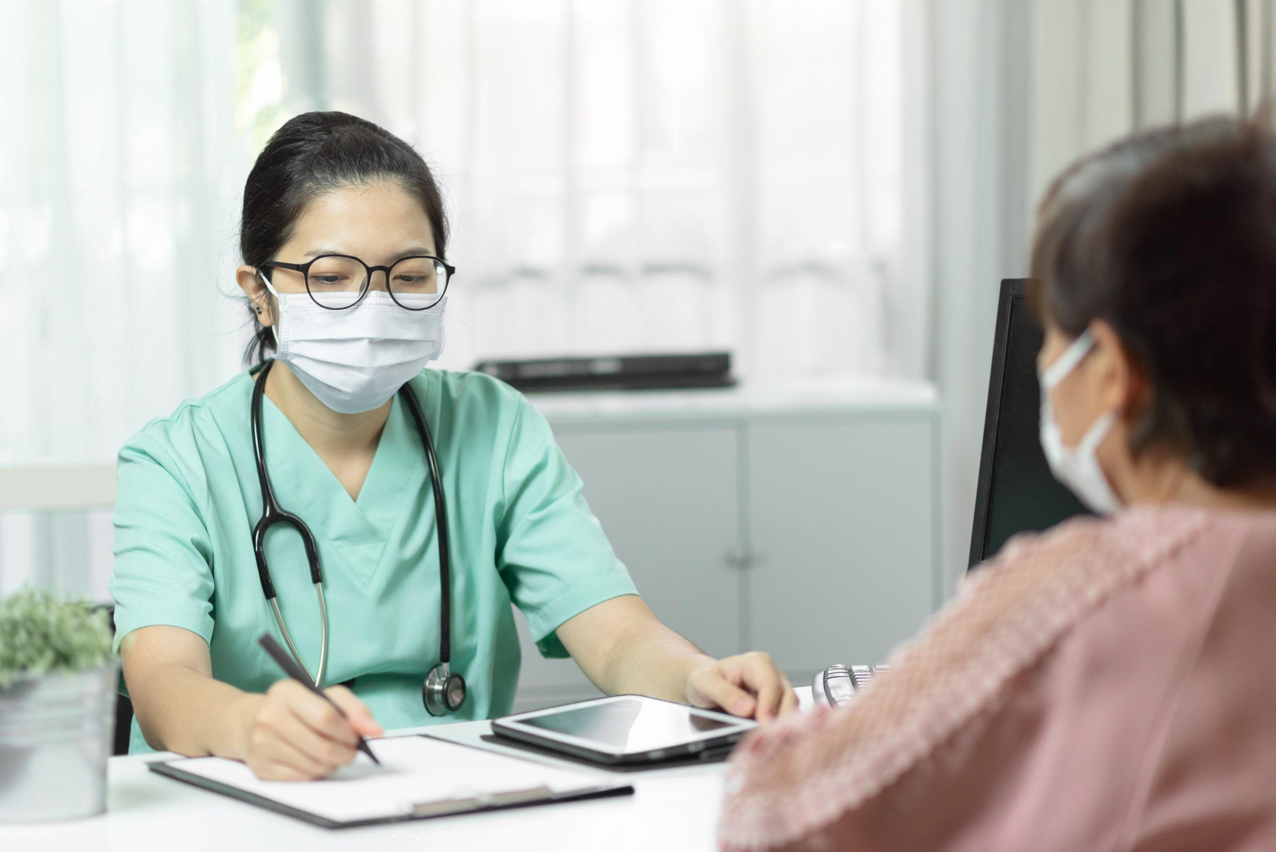Asian female Doctor in green uniform wear eyeglasses and surgical mask writing something in checklist document while talking with Elderly woman patient in medical room at the hospital. Symptoms, Disease, Flu. Covid-19, Coronavirus