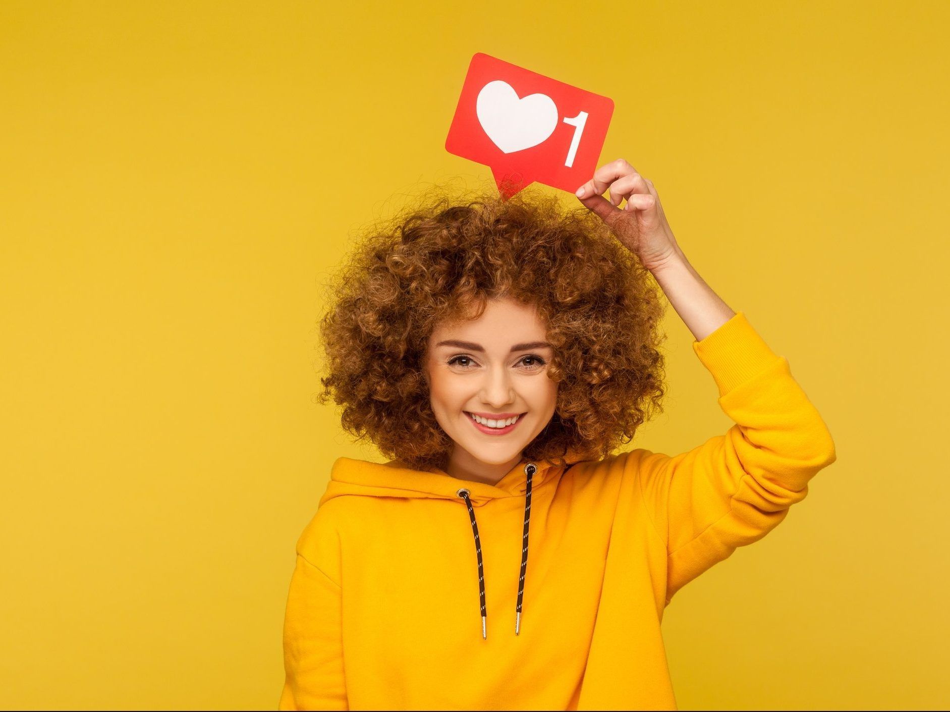Internet blogging. Portrait of happy smiling curly-haired young woman in urban style hoodie holding heart like icon