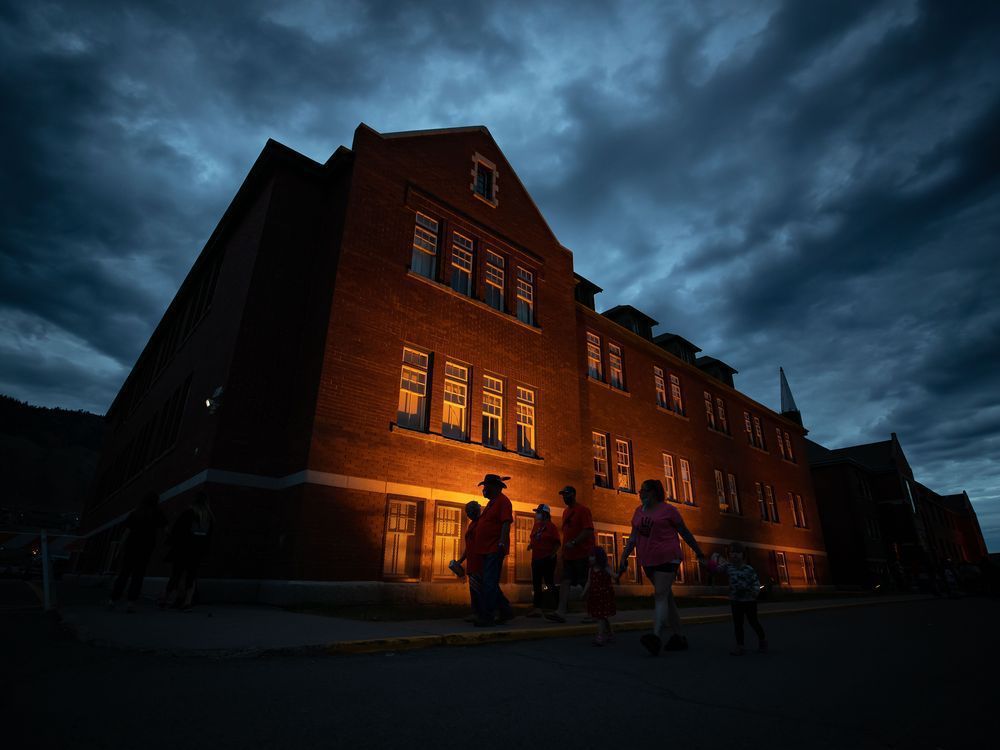 People are silhouetted as they walk past the former Kamloops Indian Residential School after gathering to honour the 215 children whose remains have been discovered buried near the facility, in Kamloops, B.C., on Monday, May 31, 2021.&nbsp;The year since the the Tk'emlups te Secwepemc First Nation announced that ground-penetrating radar had located the suspected grave sites in a former apple orchard has been one of national reckoning about residential schools in Canada.