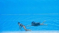 A synchronized swimmer floats motionless at the bottom of a pool, possibly due to shallow water blackout. Her coach swims behind her, reaching out to help the unconscious swimmer.