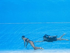 A synchronized swimmer floats motionless at the bottom of a pool, possibly due to shallow water blackout. Her coach swims behind her, reaching out to help the unconscious swimmer.