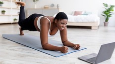 Plus size Afro woman working out at home with online personal trainer, using laptop, standing in elbow plank pose