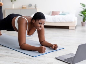 Plus size Afro woman working out at home with online personal trainer, using laptop, standing in elbow plank pose