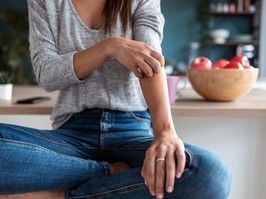 Young woman scratching her arm while sitting on the stool in the home kitchen.