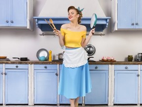 Retro pin up girl woman female housewife wearing colorful top, skirt and white apron and yellow high heels holding wooden spoons and pan standing in the blue kitchen with blue cabinets and utensils.