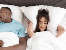 couple in bed, man is snoring, woman has pillow over ears