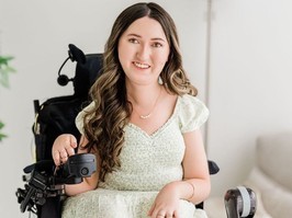 Tori Hunter, an advocate for those living with spinal muscular atrophy, sits smiling for the camera. She has long, wavy brown hair and is wearing a light green and white dress.