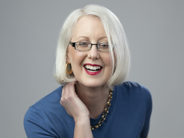 A portrait of Laura Feltz, the subject of this article and who lives with low vision from glaucoma. A woman with white-blond hair cut to a bob smiles for the camera. She is wearing black glasses and a blue shirt with a gold necklace.