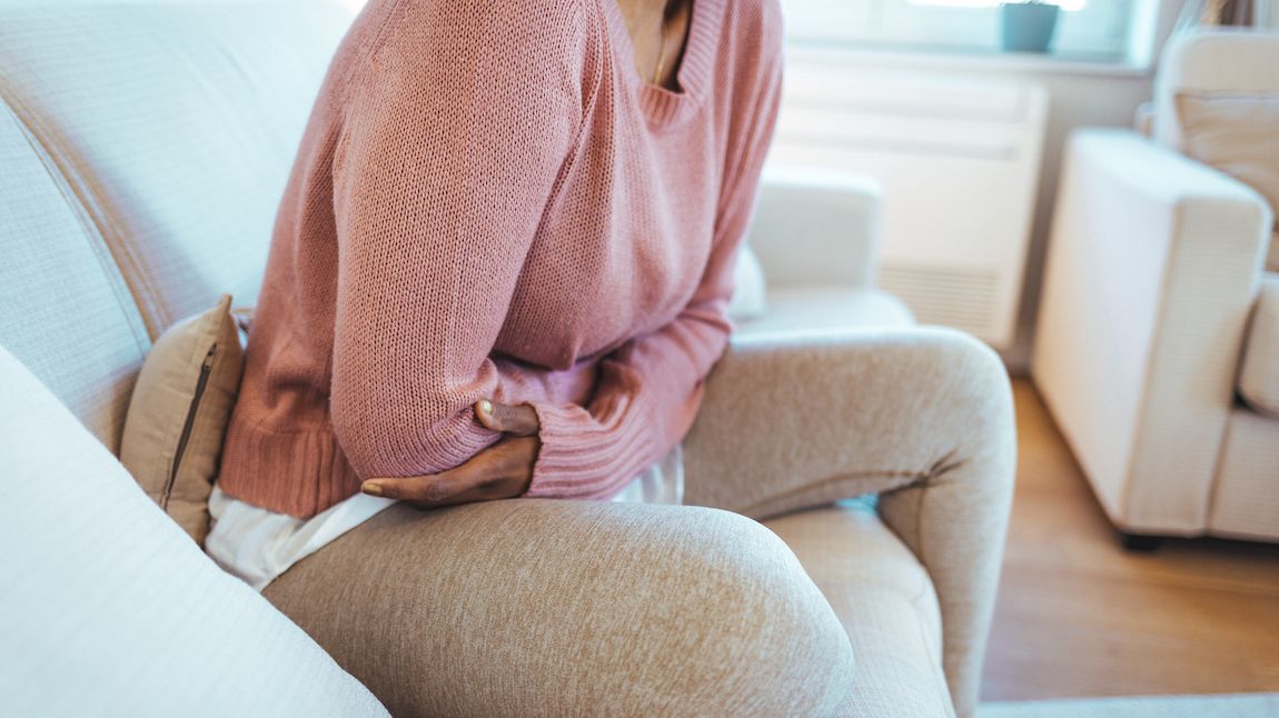 Cropped shot of a young woman holding her stomach in pain on a couch.