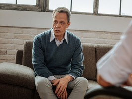 Adult man talking. Upset middle-age man sitting on grey couch in cabinet of his doctor and talking about problems in his life