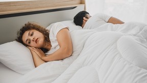 Woman and man laying back to back in bed. Woman looks upset.