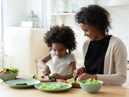 Smiling loving black mother and daughter prepare healthy delicious salad in kitchen.