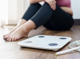 Woman sitting on the floor next to a scale and measuring tape. Upset at weight and struggle to lose weight.