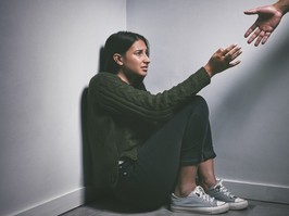 Shot of a young woman sitting in the corner of a dark room with a hand reaching out to help her - stock photo