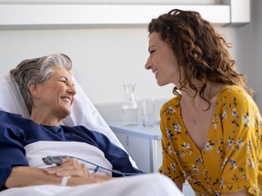 Young daughter visiting sick old mother in hospital. Happy senior patient smiling while in conversation with her lovely daughter. Granddaughter visiting and cheering her hospitalized grandmother lying in bed at hospital ward after surgery.