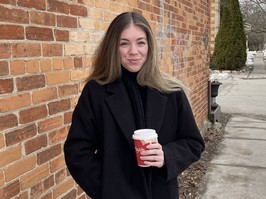 Picture of young woman Jessica Thomas standing on sidewalk next to a brick wall holding a coffee.