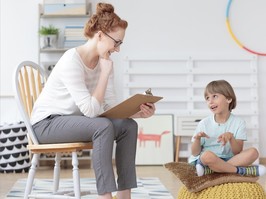 Cheerful young kid talking with helpful child counselor during psychotherapy session in children mental health center