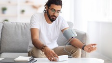 Young man sitting on couch, checking blood pressure and taking notes at home. Indian guy in white t-shirt using modern tonometer, experiencing hypertension caused by stress