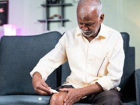 Focus on hands, Senior man applying ointment cream for joint knee pain at home while sitting on sofa - concept of treatment for osteoarthritis, knee injury and arthritis.