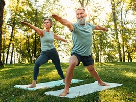 Older man and women doing yoga in the park surrounded by green grass and trees.