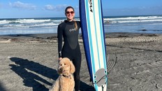 Chloe Kerton posing for a photo in a wetsuit with a surfboard and her dog. Teenage girl surfing.