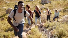 Millennial African American man leading friends hiking single file uphill on a path in countryside