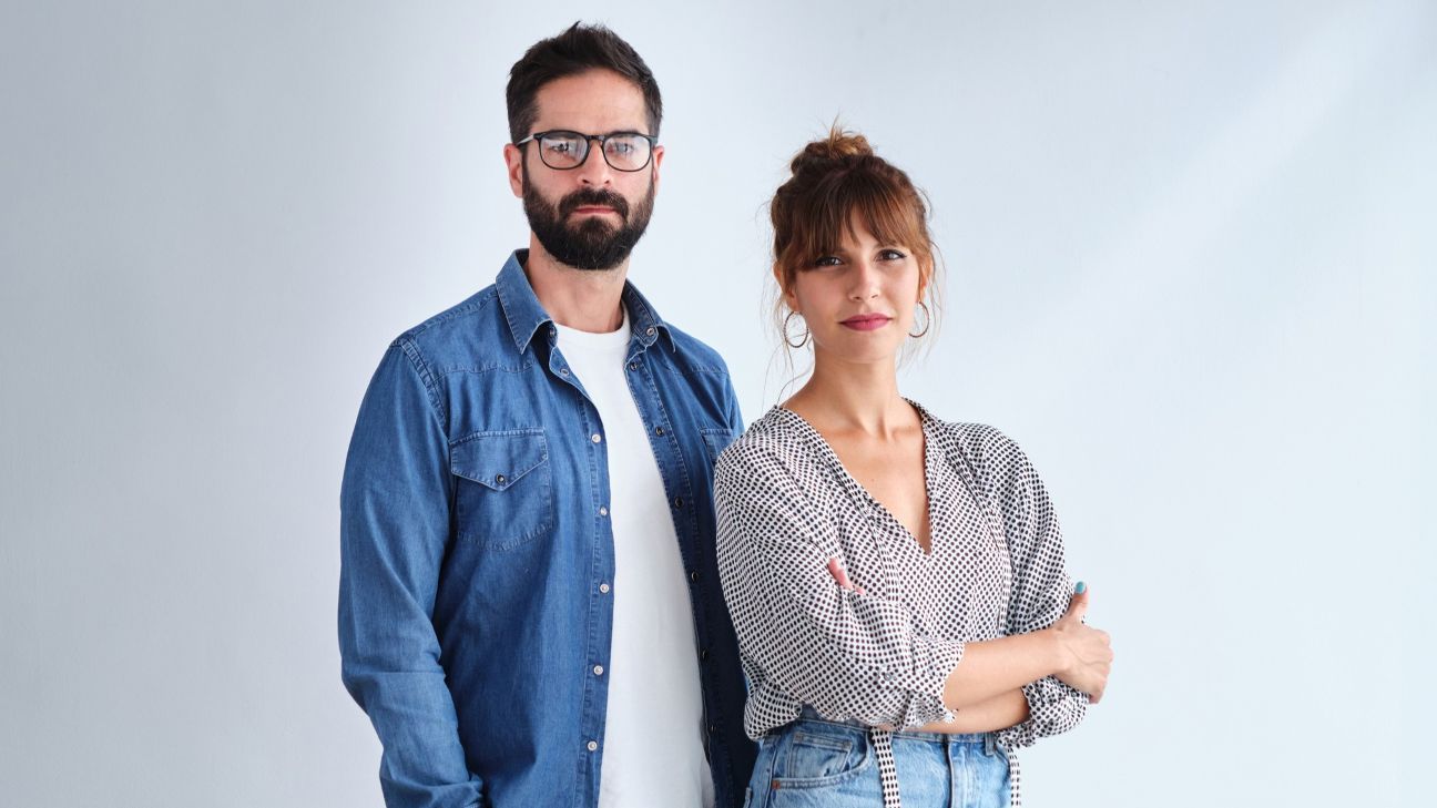 Portrait of an adult man with beard and glasses with his hands in his pants pockets and an adult woman with crossed arms looking at the camera on a white background.