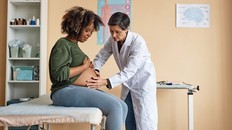 Medium long shot of pregnant young Black woman sitting on examination table while senior Caucasian obstetrician checking her bare belly with hands in hospital