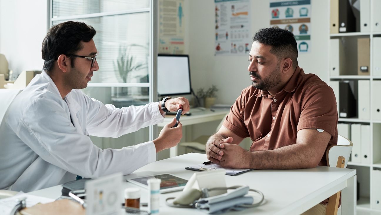 Middle aged Caucasian male doctor demonstrating glucose meter to middle aged Black man with diabetes during medical consultation in modern clinic exam room