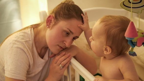 Tired mother falling asleep next to her baby son's crib.