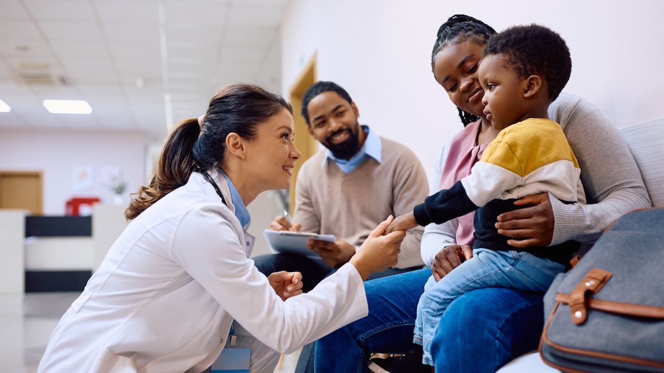 Smiling doctor holding hand on African American boy who came with parents for medical exam at pediatric clinic.