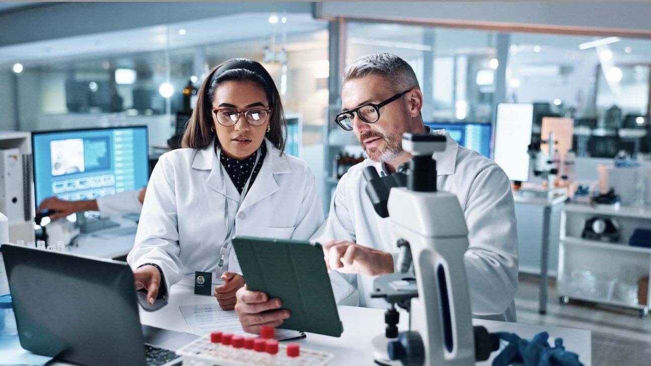 Two medical scientists work together at a desk looking over data on a tablet in a research lab.