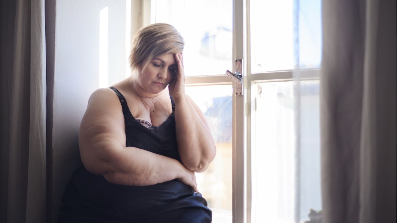 Woman living with obesity standing next to a window with her head in her hand, looking distressed.