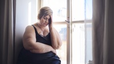 Woman living with obesity standing next to a window with her head in her hand, looking distressed.