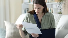 A woman in a sling reads papers on her couch with a stressed out look on her face.