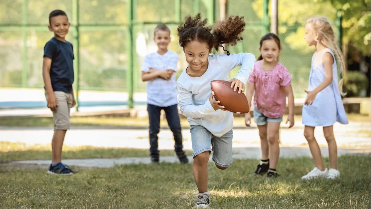 A little girl holds a football running away from her four friends in the background while they have fun together playing sports outside at a grass field park.