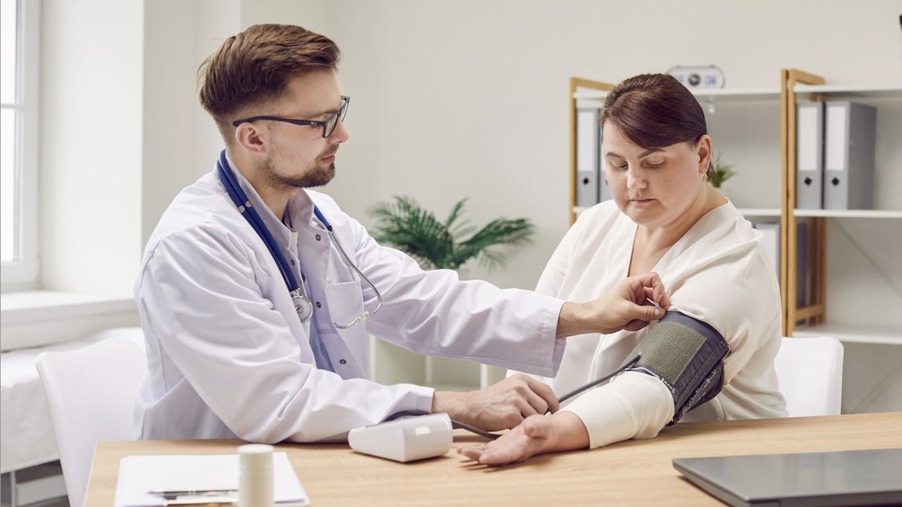 A male doctor takes the blood pressure of a female patient in a brightly lit doctor's office who lives with obesity