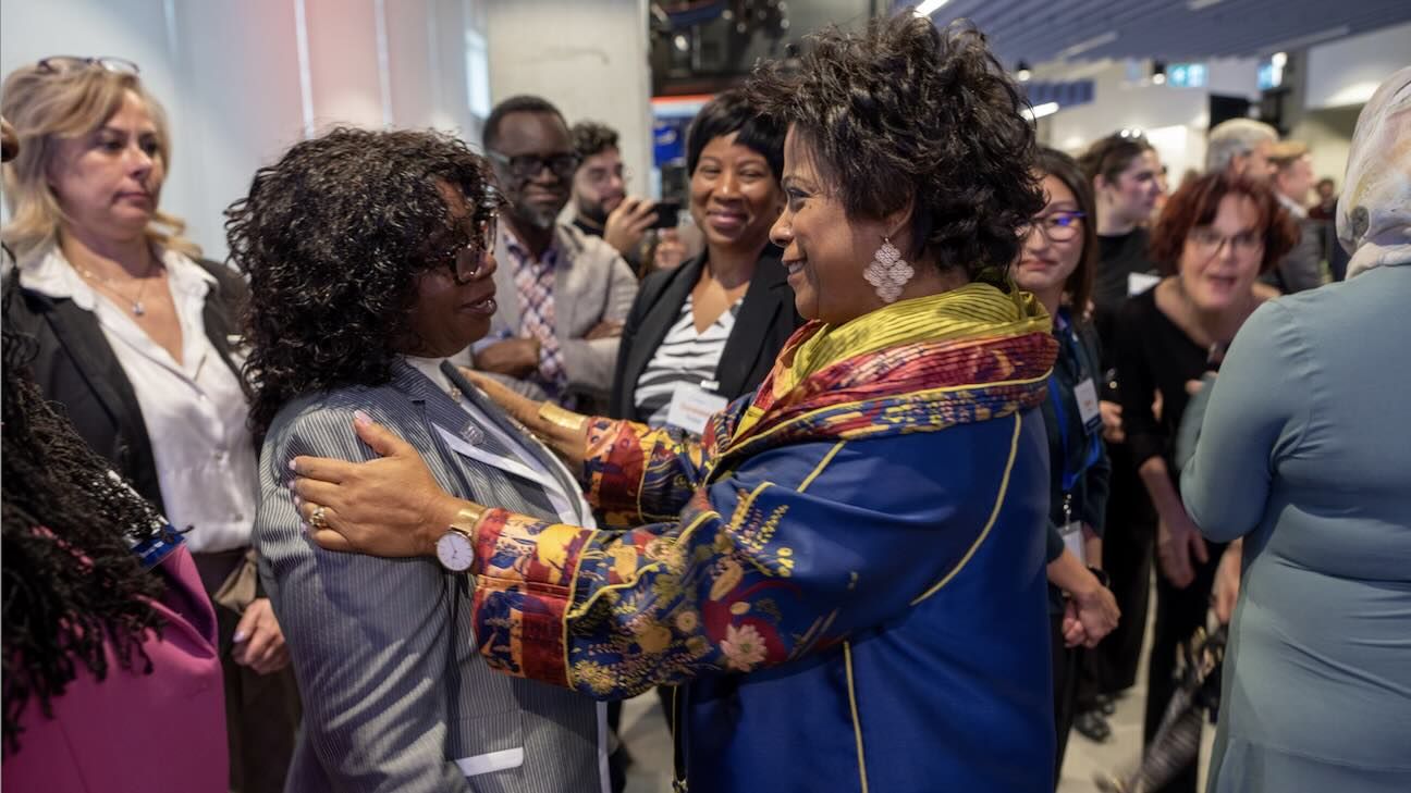 Two women standing in a crowded room at a convention with one women's hands on the other's shoulders in a warm embrace
